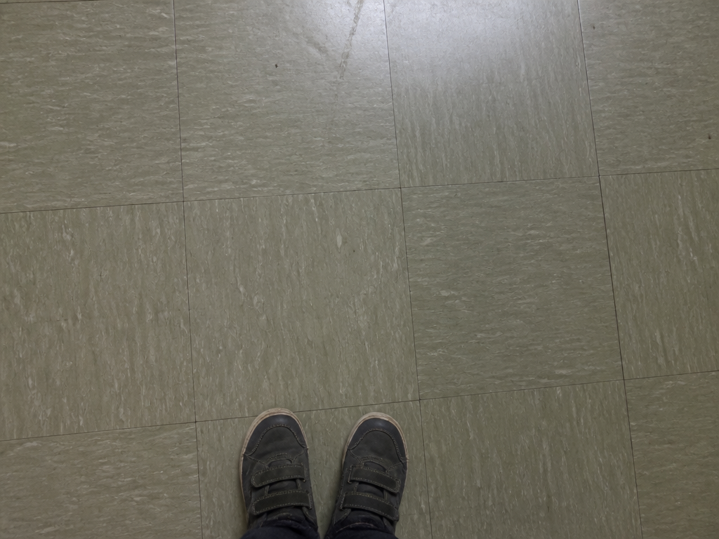 Downward view of a scuffed gray-green linoleum classroom floor with a child's small shoes at the frame edge