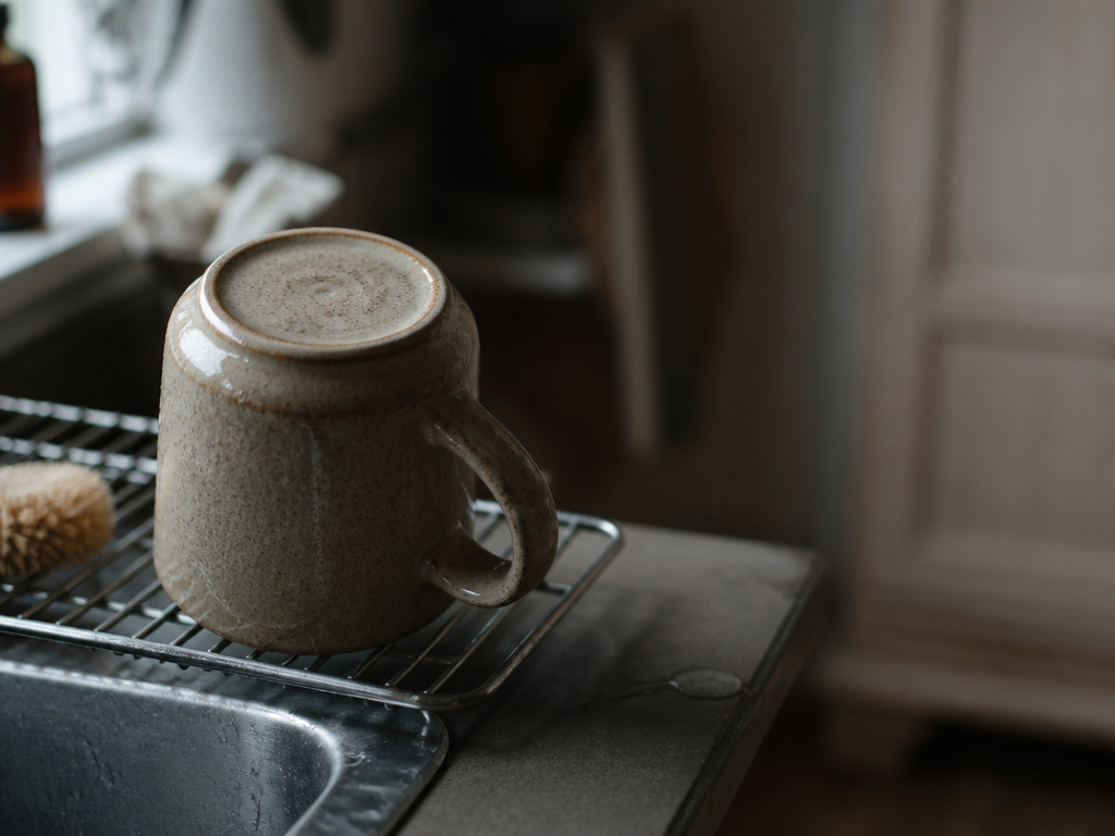 Chipped sandy-glazed stoneware mug upside down on a dish rack, water beaded on it, closed cabinet blurred behind